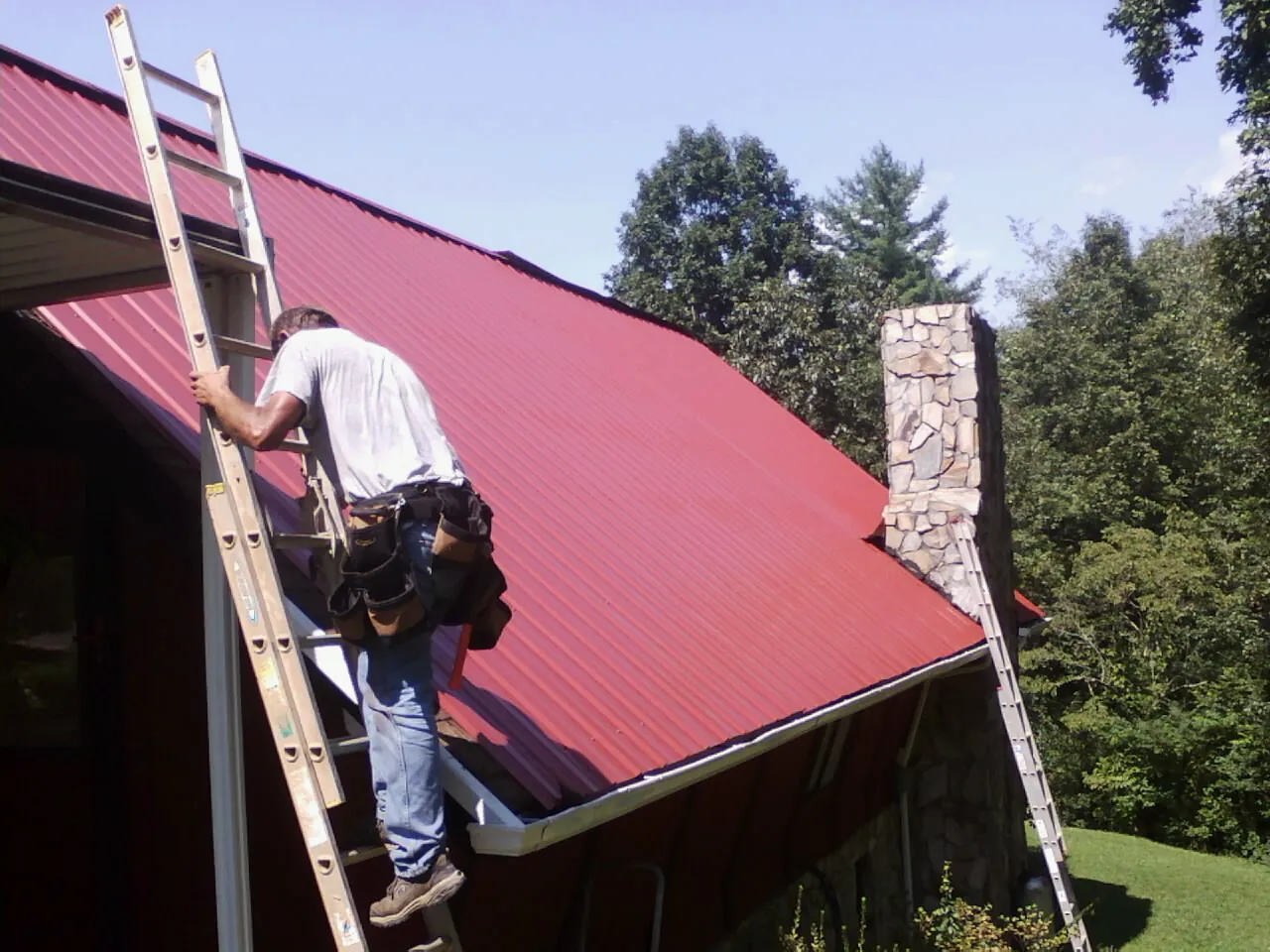 Roofer on a ladder beside a red metal roof and stone chimney — Soffit & Fascia Repair by Roofing Brothers in Saukville
